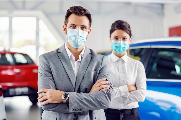 Two successful, proud car sellers standing with arms crossed in car salon and having face masks on faces