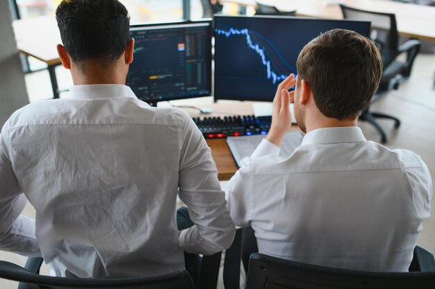 Two successful trader sitting in office and looking at screen checking cryptocurrency information data on finance market graph pointing on monitor