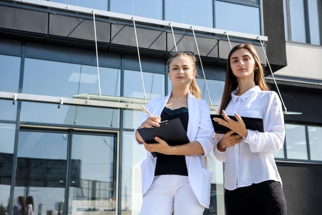 Two successful and young business ladies with tablet standing near office building