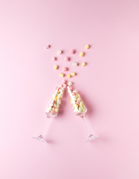 Two tall glass glasses with multicolored marshmallows clinking on a pink background. The concept of Valentine's Day.