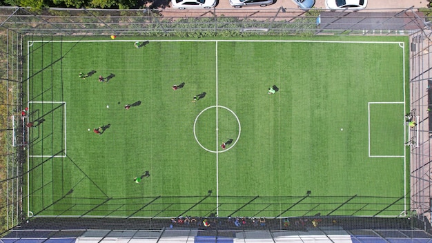 Two teams of guys are playing football on the field Top view from the drone on the ball and athletes The green field is fenced with a grid There are cars The ball hit the goal They run after ball