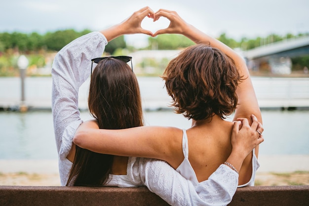 Two teenage friends making a heart shape with her hands while they are sitting on a bench by a river