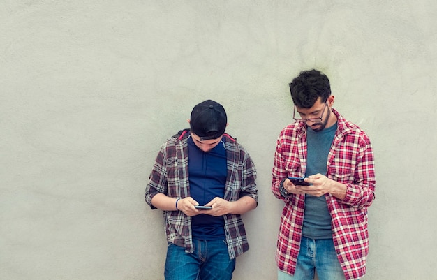 Two teenage friends on a wall checking their cell phones Two friends leaning on a wall texting on their phones Friend showing cell phone to his friend Smiling friends checking cell phones