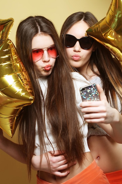 Two teenage girls friends with gold balloons make selfie on a phone Yellow background