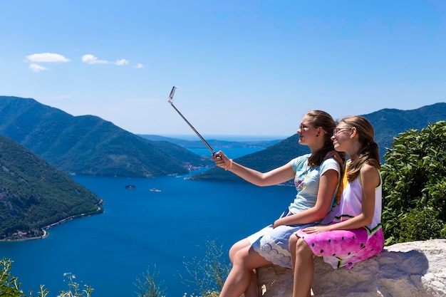 Two teenage girls in the make photo on the phone and selfie sticks on the background of Boko Kotorska bay and mountains in Montenegro