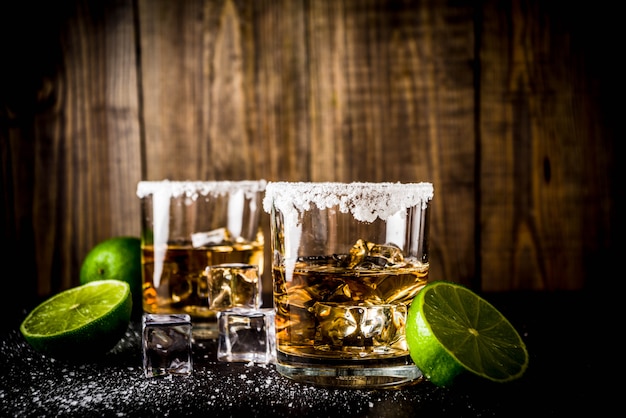 Two tequila shot glasses on dark table, with ice cubes, salt and limes