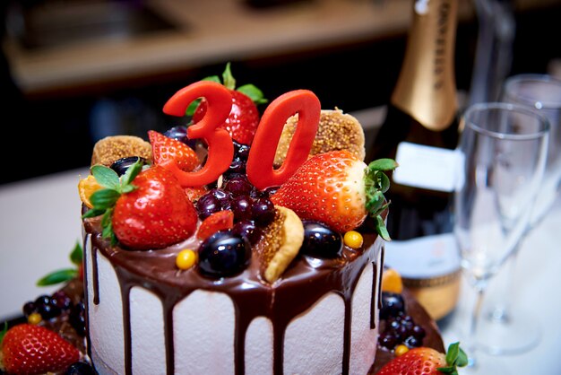 A two-tiered white cake with fresh fruit and chocolate stands next to a bottle of champagne 