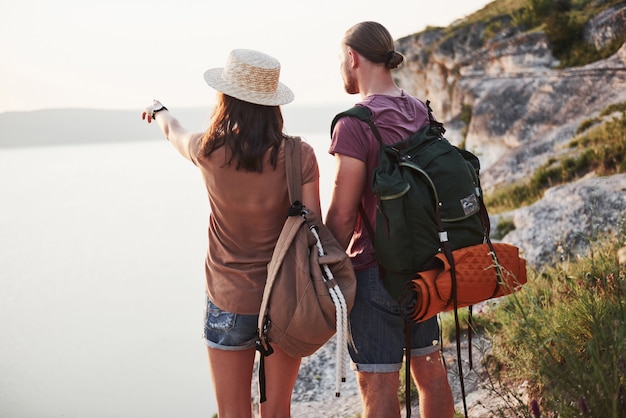 Two tourist male and woman with backpacks stand to the top of the crag and enjoying sunrise. Traveling mountains and coast, freedom and active lifestyle concept