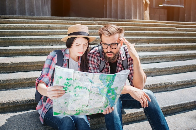 Two tourists sitting on steps and hold map together. They look at it. People are concentrated and serious.