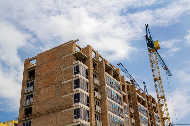 Two tower cranes working on high rise brick building under construction on blue sunny sky copy space background. Modern urbane architecture, investments, buying and selling concept.