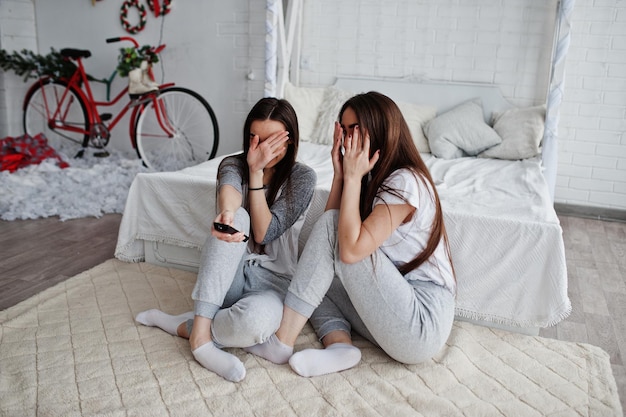 Two twins models girls while click the remote control from the TV on studio and hiding faces becouse they watched scary movie They are frightened and shocked