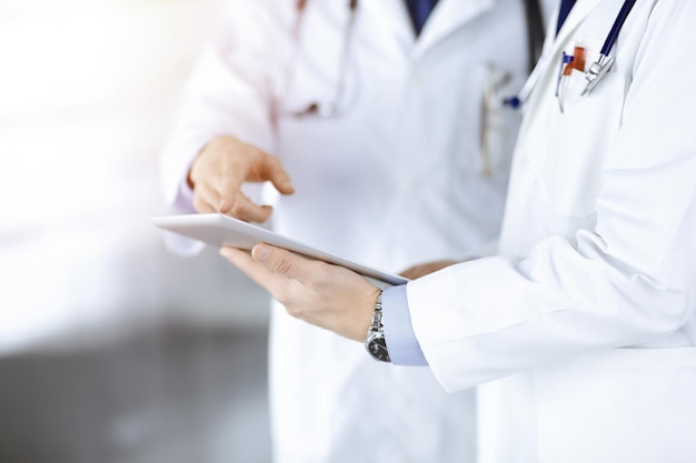 Two unknown doctors with stethoscopes discussing medical exam resoults, standing at sunny hospital office. Physicians use a computer tablet for filling up medication names records. Perfect medical ser