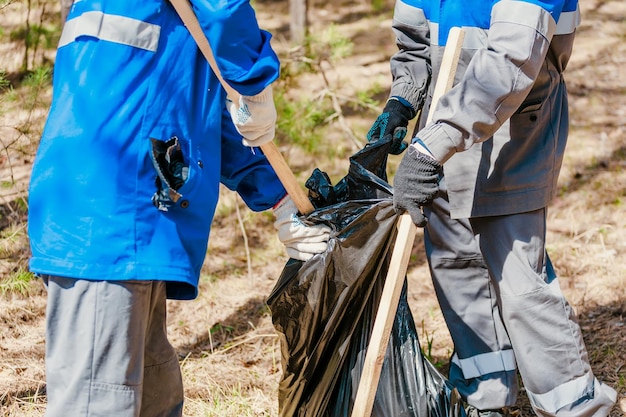 Two volunteers in work overalls and gloves collect garbage in forest in plastic bag Closeup during action