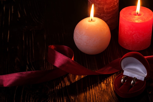 Two wedding rings in red gift box with three wax flame candlelight with ribbon in dark romantic light, love dating, Valentine's day, selective focus