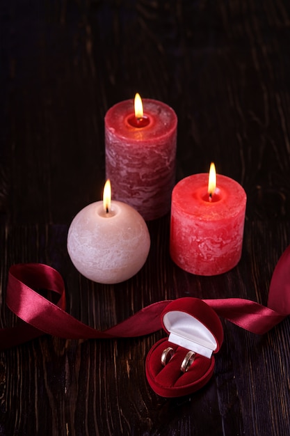 Two wedding rings in red gift box with three wax flame candlelight with ribbon in dark romantic light, love dating, Valentine's day, selective focus