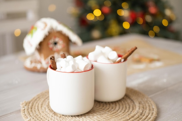 two white cup of cocoa with marshmallows and gingerbread house on table