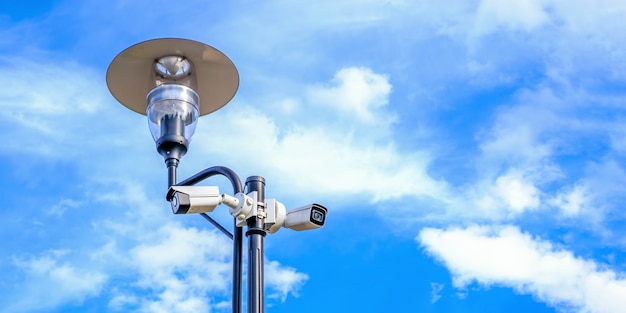 Two white surveillance cameras on the metal street light lamp post outdoor on blue sky