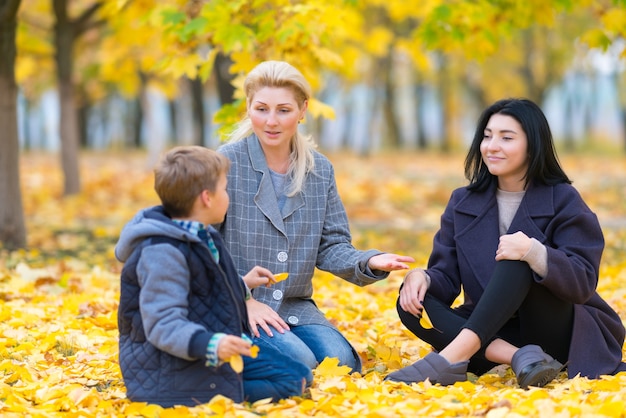 Two woman and teenage boy in park sitting on the ground covered with bright yellow leaves and chatting together