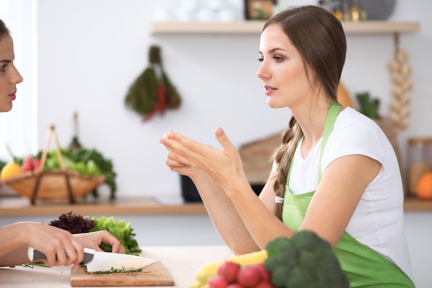 Two women are cooking fresh salad in a kitchen and having a pleasure talk Friends and Chef Cook concept