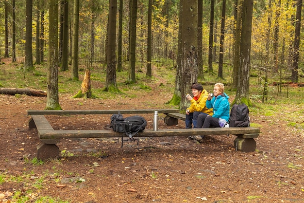 Two women are laughing, sitting on a bench in forest Drink tea or coffee from a thermos