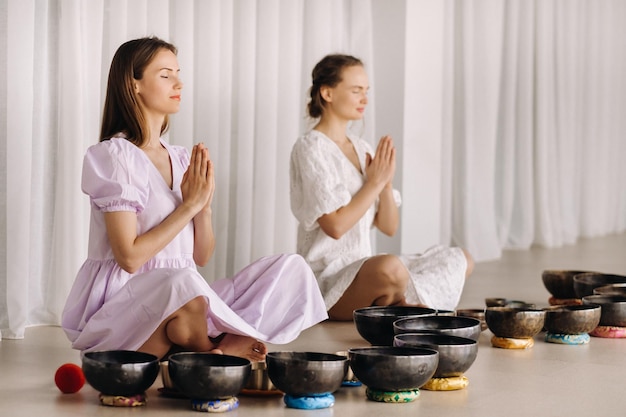 Two women are sitting with Tibetan bowls in the lotus position before a yoga class in the gym