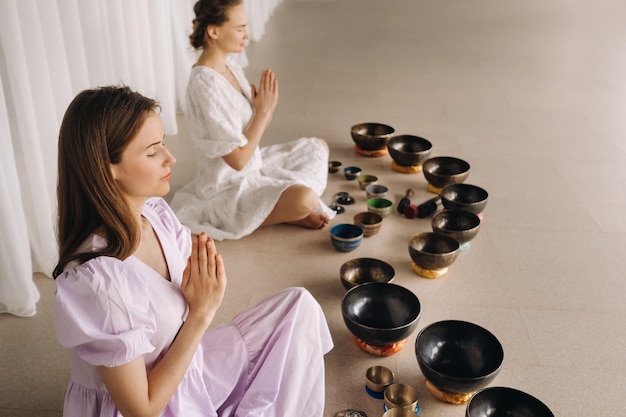 Two women are sitting with Tibetan bowls in the lotus position before a yoga class in the gym