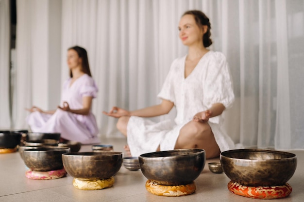 Two women are sitting with Tibetan bowls in the lotus position before a yoga class in the gym