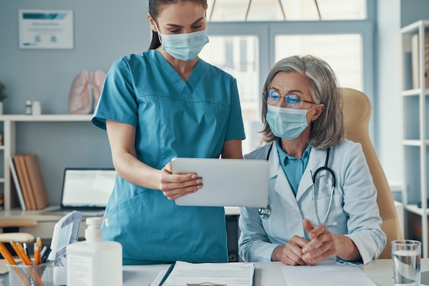 Two women coworkers in medical uniform and protective masks talking and using digital tablet while working in the hospital