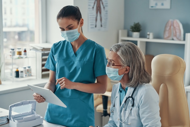 Two women coworkers in medical uniform and protective masks talking and using digital tablet while working in the hospital