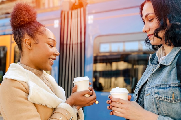 Two women in a denim jacket talking to each other laughing drinking coffee and waiting for a tram