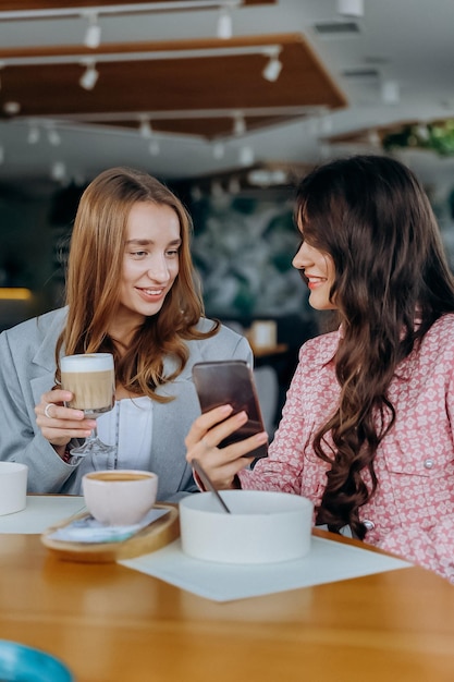 Two women drinking coffee while sitting in cafe or restaurant and looking on mobile phone