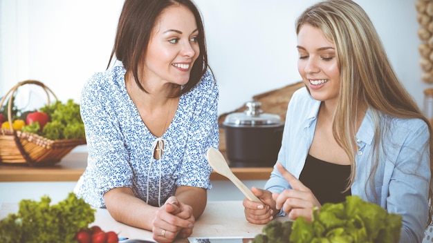 Two women friends choosing the recipe for a delicious meal while sitting at the table in the kitchen Tablet pc is the best cookbook