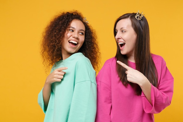 Two women friends european african american girls in pink green clothes, crowns posing isolated on yellow background. People lifestyle concept. Mock up copy space. Point index fingers at each other.