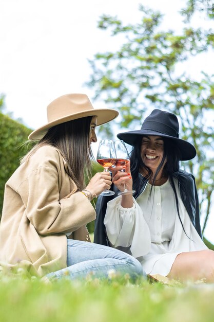 Two women friends in hats sitting on the grass toasting with glasses of wine Lifestyle of young friends in hats sitting on the grass toasting with wine