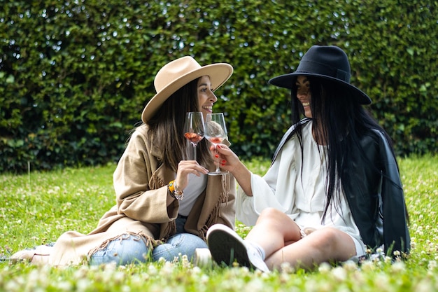 Two women friends in hats sitting on the grass toasting with glasses of wine.