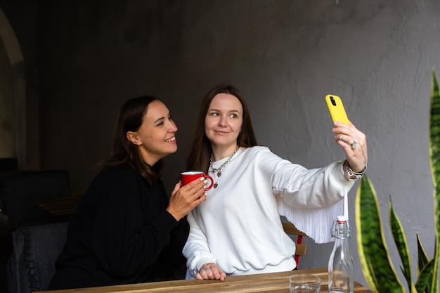 Two women friends have fun outdoors in cafe and take a selfie on a smartphone