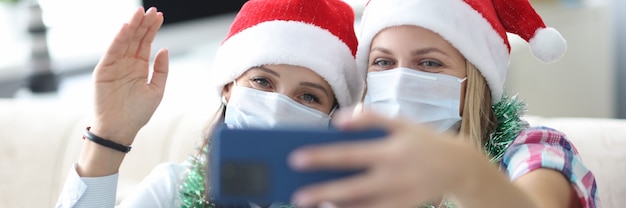 Two women friends in protective face masks and red santa hats are photographed on phone portrait