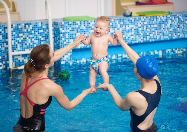 Two women holding happy laughing baby doing exercising over water in air during swimming classes Baby relaxing in pool