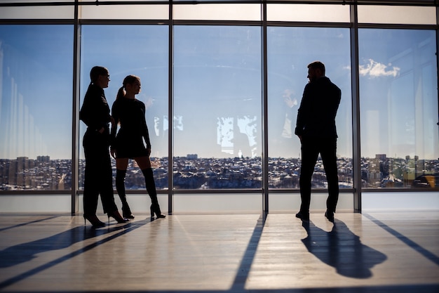 Two women and a man in the office enjoy the view of the city from the window of a skyscraper and talk while standing by the large window in the office