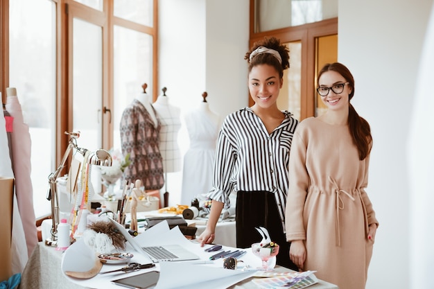 Two women stylists in design studio. Work process of modeling new clothes. African and caucasian spectacled women are looking to the camera