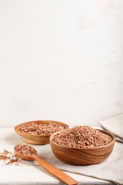 Two wooden bowls with unpolished brown rice and wooden spoon on a white wooden background. Side view, copy space.