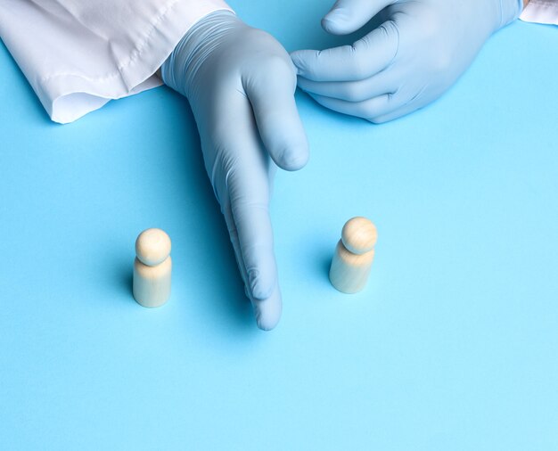 Two wooden figures of men stand against the background of the male hands of the doctor in blue latex gloves, keep a distance of one and a half meters during the epidemic and pandemic