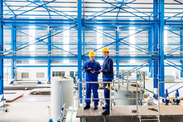 Two workers in large metal workshop or factory checking work standing on large machine