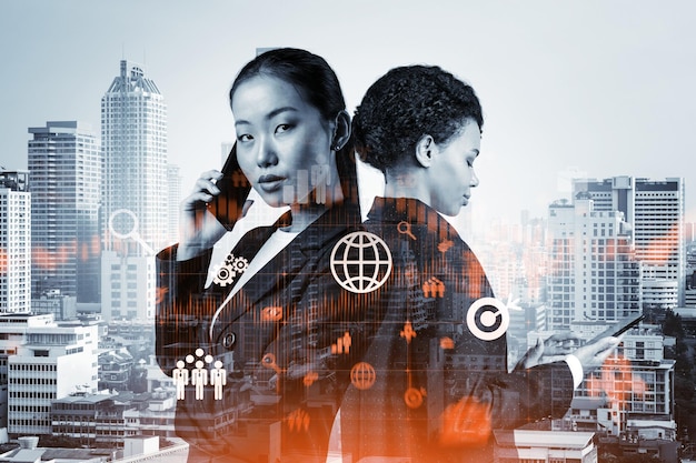 Two young astonishing businesswoman in suits try to find new tech approaches to solve problems at research and development department Fintech hologram icons over Bangkok background