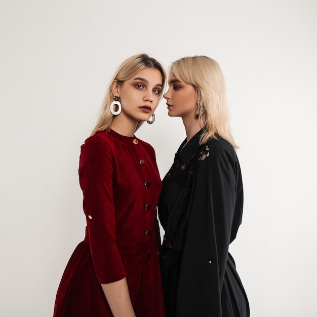 Two young attractive blonde women in fashionable clothes stand near white vintage wall