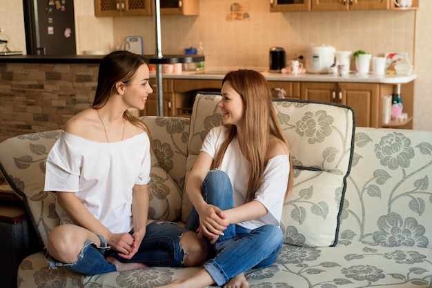 Two young beautiful cheerful girlfriends in blue jeans sitting on sofa at home laughing.