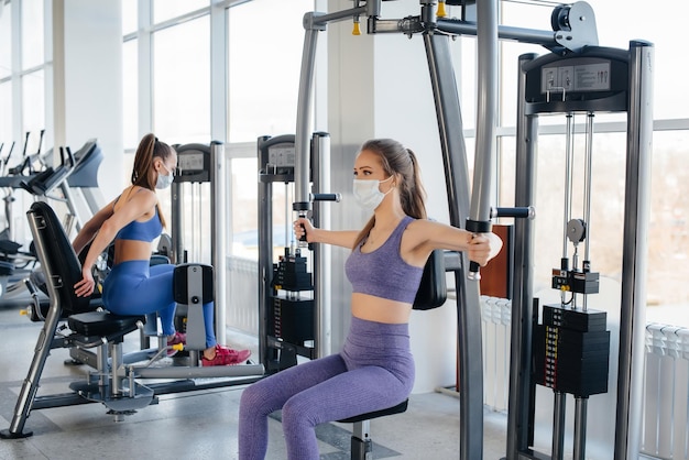 Two young beautiful girls exercise in the gym wearing masks during the pandemic.