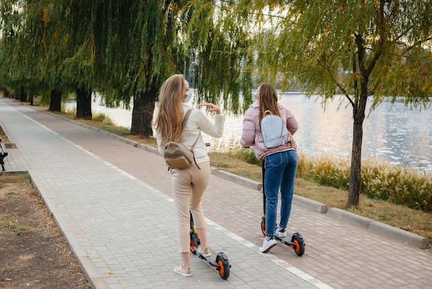 Two young beautiful girls in masks ride electric scooters in the Park on a warm autumn day. Walk in the Park.