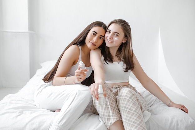 Two young beautiful girls with cosmetics sit on a white bed Women use serum cream and facial lotion to take care of their skin Girlfriends in home spa rest and relax