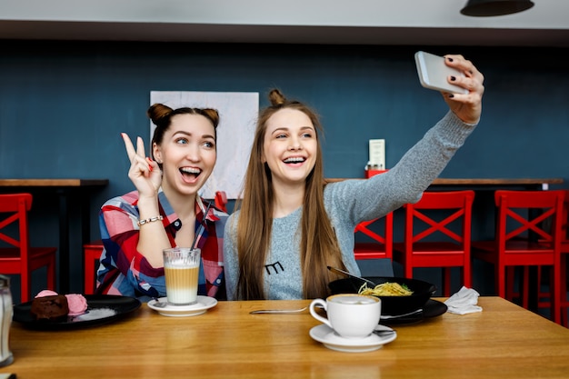 Two young beautiful hipster women sitting at cafe, stylish trendy outfit, europe vacation, street style, happy, having fun, smiling, sunglasses, looking at smartphone, taking selfie photo, flirty